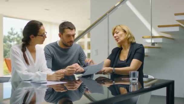 Assis à la table dans le salon agent immobilier et beau jeune couple signe contrat et achète la maison. La maison est ensoleillée et lumineuse. Le couple est heureux, tout le monde sourit .