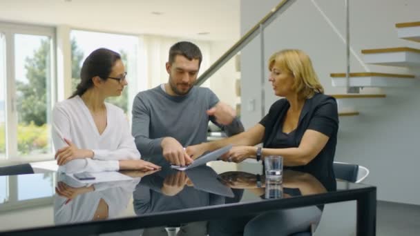 Assis à la table dans le salon agent immobilier et beau jeune couple signe des documents et heureusement embrasser. La maison est ensoleillée et lumineuse .