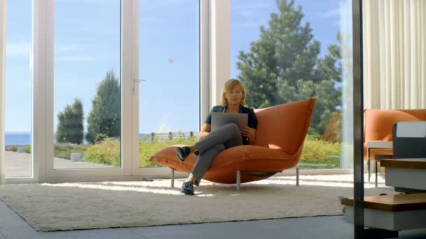 高级的女人使用笔记本电脑坐在她阳光的客厅里的沙发上。房子有地板到天花板的窗户与拥有视图。天气晴朗. — 图库视频影像