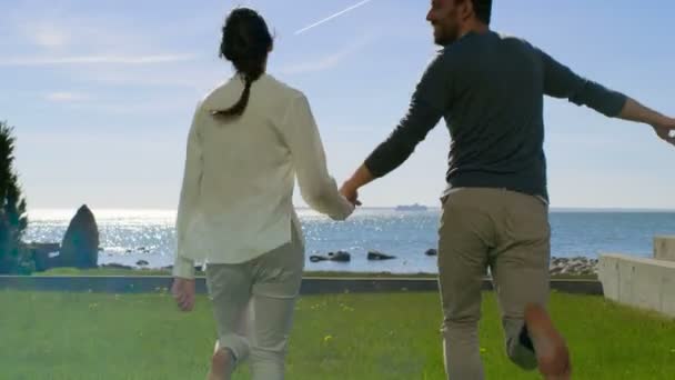 En el hermoso día de verano Pareja feliz cogida de la mano y correr descalzo sobre la hierba. Antes de ellos Sunny Blue Sky y vista al mar . — Vídeo de stock