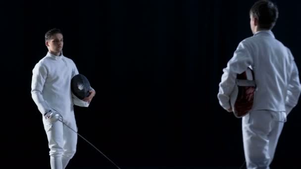 Due giovani schermidori salutano il pubblico e l'un l'altro, indossano maschere e iniziano a combattere la spada con le lamine. Girato sullo sfondo nero isolato . — Video Stock