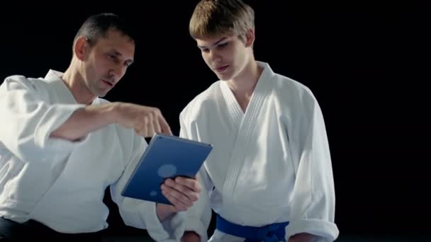 Kampfsportlehrer mit Hakamas bringen jungen Studenten Aikido-Technik mit Hilfe des Tablet-Computers bei. Aufnahme isoliert auf schwarzem Hintergrund und in Zeitlupe. — Stockvideo