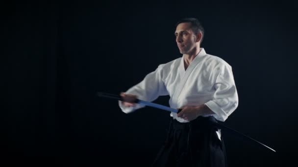 Portraitaufnahme des Aikido-Meisters in traditioneller Samurai-Hakama-Kleidung, der sein japanisches Schwert aus der Scheide holt und damit schwingt. Er steht im Rampenlicht, Dunkelheit umgibt ihn. Schuss isoliert auf schwarzem Hintergrund. — Stockvideo