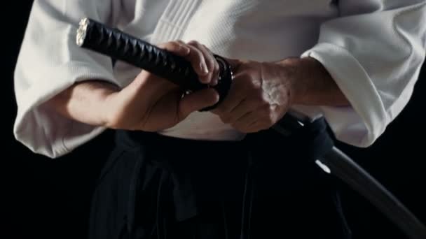 Aikido Master dragen traditionele Samurai Hakama kleding neemt zijn Japans zwaard uit de schede en schommels met het. Hij is in de Spotlight duisternis omringt hem. Schot geïsoleerd op zwarte achtergrond. — Stockvideo