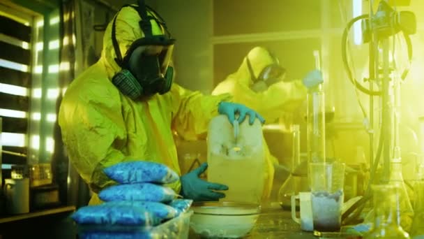 In het laboratorium van de ondergrondse Drug mengen clandestiene chemici dragen van beschermende maskers en overall chemicaliën. Men giet vloeistof uit de bus in Bowl, tweede controles bekerglas voor Product consistentie. Ze werken in de verlaten gebouw. — Stockvideo