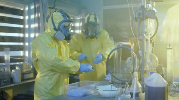 Dans le laboratoire souterrain, deux chimistes clandestins portant des masques de protection et des combinaisons emballent des sacs remplis de méthamphétamine cristalline dans des boîtes. Le laboratoire est plein d'équipements illégaux. Ils s'accroupissent dans un bâtiment abandonné . — Video