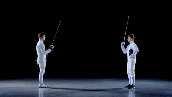 Two Young Professional Fencers Greet Each Other, and Preparing for Fighting Match. Shot Isolated on Black Background.