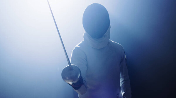 Полностью экипированный "Фенкер" поднимает меч из фольги в готовности к матчу. Он стоит в центре внимания, в то время как тьма вокруг Него. Выстрел на черном фоне
.