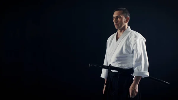 Retrato Shot of the Aikido Mestre vestindo roupas tradicionais Samurai Hakama detém sua espada japonesa. Ele está no Spotlight Darkness rodeia-o. Tiro isolado no fundo preto . — Fotografia de Stock
