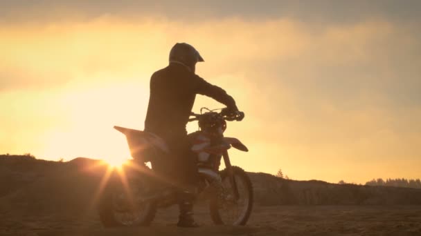 Professionele Fmx motorrijder berust op zijn fiets en kijkt uit op harde Sandy Off-Road terrein. Zon ondergaat. — Stockvideo