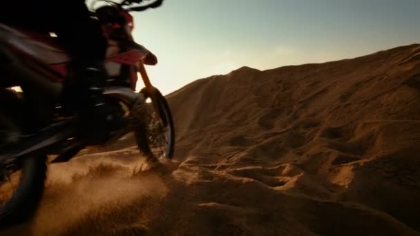 Ángulo bajo que sigue al tiro del motociclista profesional que conduce sobre la duna y más abajo en la pista todoterreno. Es puesta del sol y la pista está cubierta con humo / niebla . — Vídeo de stock