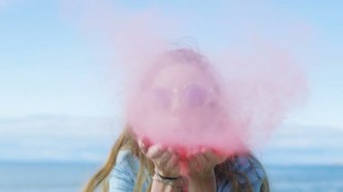 Güzel genç genç kız kahverengi saçlı ve serin güneş gözlüğü elleri ve gülüyor Holi renkli pudrayı darbeler. Onun arkasında açık mavi gökyüzü.