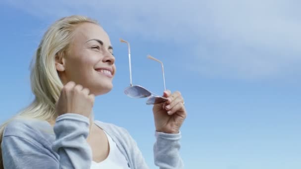 Douce jeune blonde court la main à travers ses beaux cheveux blonds et mettre sur Aviator Lunettes de soleil. Elle regarde loin dans le ciel clair lumineux et l'avenir sans nuages . — Video