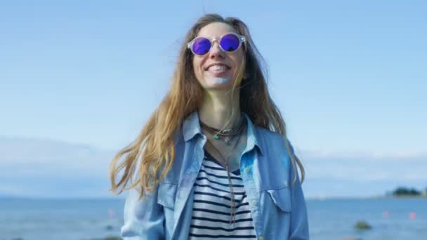 Jovem menina bonita com cabelos castanhos vestindo óculos de sol frescos joga Holi pó colorido para o ar e ri. Céu azul claro atrás dela e mar azul profundo atrás dela . — Vídeo de Stock