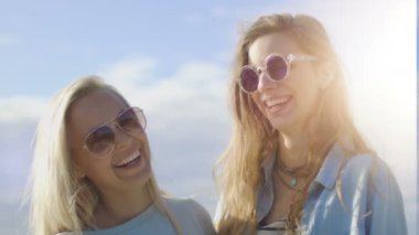 Portre çekim iki güzel mutlu kız sarılma, poz ve kameraya gülümseyen güneş gözlüğü takıyor. Yaz eğlenceli var. Açık mavi gökyüzü arka planda görülür. 