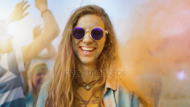 Närbild porträtt av en vacker ung flicka med solglasögon står i mängden av människor fira Holi festivalen. Människor kastar färgglada pulver i ryggen. Hennes ansikte och kläder är täckta med färgglada pulver. — Stockvideo
