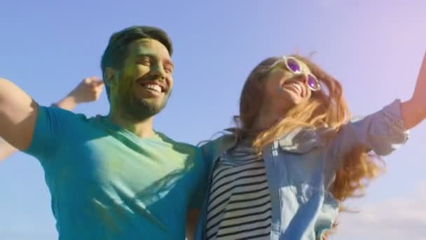 Retrato de una feliz pareja cubierta de colorido polvo bailando en el festival Holi. La gente en el fondo se divierten tirar Holi polvo . — Vídeos de Stock