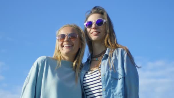 Dos hermosas chicas felices que usan gafas de sol abrazan, ríen y se divierten mucho. Cielo azul claro en el fondo . — Vídeo de stock