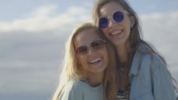 戴着太阳镜，拥抱、 合影和微笑在相机上的两个美丽快乐女声的肖像镜头。他们有很多夏天的乐趣。在后台看到晴朗的蓝色天空. — 图库视频影像