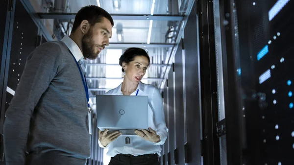 Mannelijke Server ingenieur kijkt naar Server Rack behuizing. Zijn vrouwelijke collega werkt op een Laptop naast hem. Ze zijn in grote datacenter. — Stockfoto