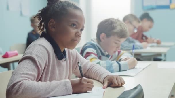 Στην τάξη του δημοτικού σχολείου λαμπρό μαύρο κορίτσι γράφει σε τετράδιο άσκηση, λαμβάνοντας δοκιμή και γράφοντας εξετάσεις. Junior Classroom with Diverse Group of Children Working επιδέξια και Μαθαίνοντας Νέα πράγματα — Αρχείο Βίντεο