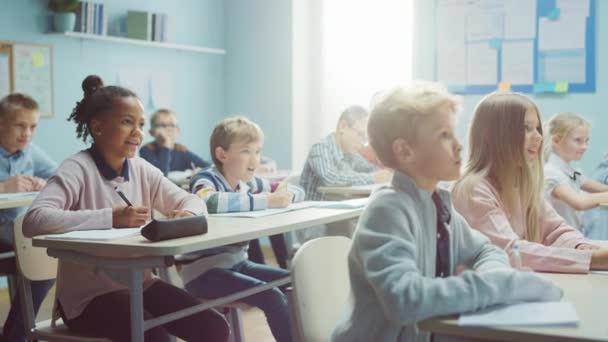 Elementary Classroom of Diverse Bright Children Listening to their Teacher Giving Lesson, Skratta tillsammans efter att han säger något roligt. Glada unga barn i grundskolan skriver i övningsanteckningar — Stockvideo