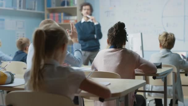 Uważny nauczyciel wyjaśnia lekcję w klasie pełnej jasnych, zróżnicowanych dzieci. W szkole podstawowej z grupą jasnych wieloetnicznych dzieci nauka uczenia się, Kid podnosi rękę z odpowiedzią — Wideo stockowe