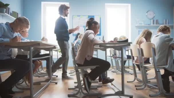 En la clase de la escuela primaria: El maestro entusiasta camina entre filas de niños brillantes y diversos, explica la lección. Grupo de niños multiétnicos inteligentes que aprenden cosas nuevas. Vista lateral Tracking Shot — Vídeos de Stock