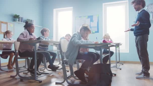 Enthousiaste Leraar legt les uit aan een klaslokaal vol diverse kinderen. In Elementary School Group of Smart Multiethnic Kids Writing in Exercise Notebooks and Learning. Zijaanzicht bewegend schot — Stockvideo