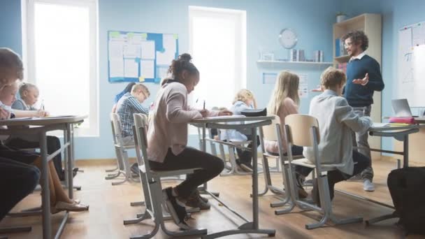 Enthousiaste Leraar legt les uit aan een klaslokaal vol diverse kinderen. In Elementary School Group of Smart Multiethnic Kids Writing in Exercise Notebooks and Learning. Zijaanzicht bewegend schot — Stockvideo