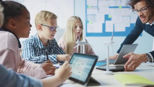 Salle de classe de l "école primaire : un enseignant enthousiaste tient une tablette d'ordinateur explique à un jeune enfant brillant comment fonctionnent les éoliennes. Les enfants s'initient aux formes d'énergie renouvelable respectueuses de l'environnement — Video