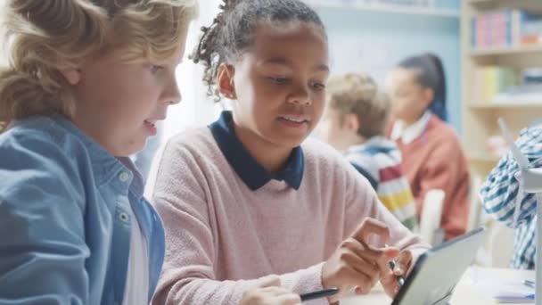 En la Escuela Primaria: Clase de Niños Pequeños Inteligentes Trabajan en Equipo Usando Computadoras Tablet para Programar Turbinas Eólicas. Aula con niños Aprendiendo sobre formas ecológicas de energía renovable — Vídeo de stock