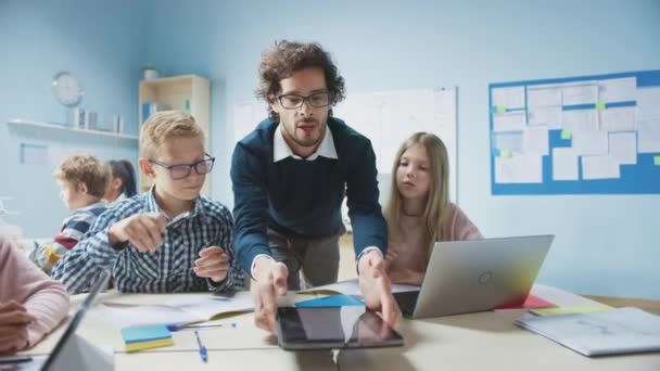 Aula de la escuela primaria: El entusiasta profesor que sostiene la computadora de la tableta explica la lección a los niños pequeños brillantes. Niños Aprendizaje Programación Idiomas, Seguridad en Internet y Tecnología de la Información — Vídeo de stock