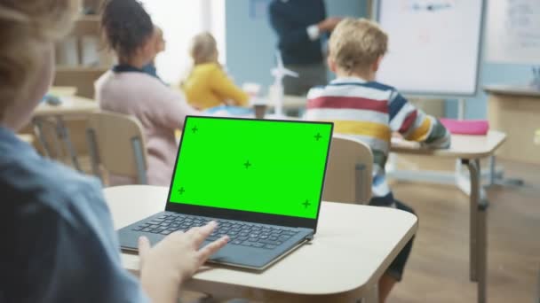 Aula de la escuela primaria: sobre el hombro vista de un niño usando Green Mock-up Screen Laptop en clase. En el fondo, el maestro explica la lección a la clase diversa — Vídeos de Stock