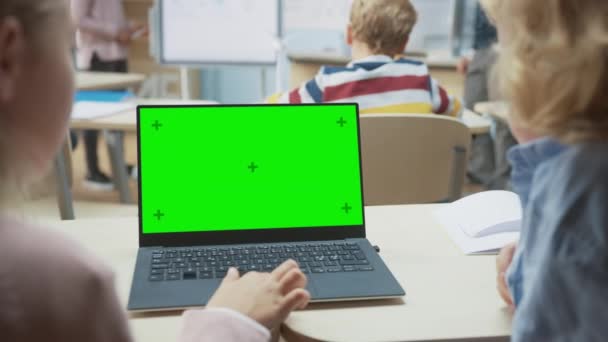 Salle de classe de l'école élémentaire : vue sur l'épaule de deux enfants à l'aide d'un ordinateur portable à écran vert maquillé en classe. En arrière-plan L'enseignant explique la leçon à donner à une classe diversifiée — Video