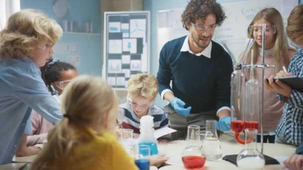Učebna základních věd: Nadšený učitel vysvětluje chemii různorodé skupině dětí, ukazuje jim, jak míchat chemikálie v kádinkách. Děti používají digitální tablety a mluvit — Stock video
