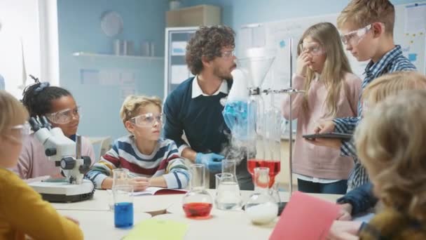 Grundskola Science Classroom: Entusiastisk lärare förklarar kemi för olika grupper av barn, visar dem hur man blandar kemikalier i bägare. Barn använder digitala surfplattor och pratar — Stockvideo