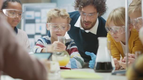 Grundskola Science Classroom: Entusiastisk lärare förklarar kemi för olika grupper av barn, små pojke blandar kemikalier i bägare. Barn lär sig med intresse — Stockvideo