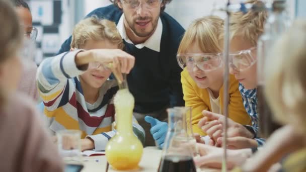 Класс естественных наук начальной школы: Энтузиастичный учитель объясняет химию различным группам детей, маленький мальчик смешивает химикаты в стаканах. Дети учатся с интересом — стоковое видео