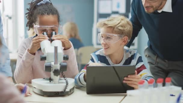 Δημοτικό Σχολείο Science Classroom: χαριτωμένο κοριτσάκι φαίνεται κάτω από το μικροσκόπιο, αγόρι χρησιμοποιεί ψηφιακό Tablet υπολογιστή για να ελέγξετε τις πληροφορίες στο Διαδίκτυο, ενώ Ενθουσιαστικός δάσκαλος εξηγεί Μάθημα — Αρχείο Βίντεο