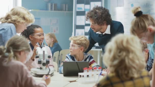 Základní škola věda učebna: roztomilé holčička vypadá pod mikroskopem, chlapec používá digitální tablet počítač pro kontrolu informací na internetu a dělat High Five v oslavě. Učitel pozoruje — Stock video