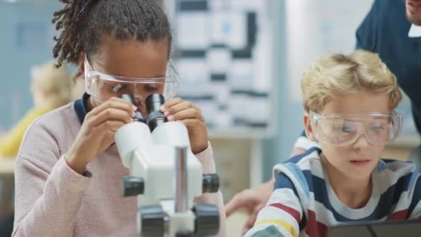 Δημοτικό Σχολείο Science Classroom: χαριτωμένο κοριτσάκι φαίνεται κάτω από το μικροσκόπιο, Αγόρι Χρησιμοποιεί Ψηφιακό Tablet υπολογιστή για να ελέγξετε τις πληροφορίες στο Διαδίκτυο. Ο δάσκαλος παρατηρεί από πίσω — Αρχείο Βίντεο