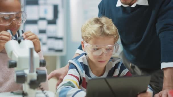 Elementary School Science Classroom: Nettes kleines Mädchen sieht unter dem Mikroskop, Junge verwendet digitale Tablet-Computer, um Informationen im Internet zu überprüfen. Lehrer beobachtet von hinten — Stockvideo