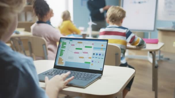 Classe de science de l'école primaire : Sur l'épaule, le petit garçon utilise un ordinateur portable avec un écran montrant un logiciel de programmation. Un professeur de physique explique la leçon à une classe diversifiée remplie d'enfants intelligents — Video