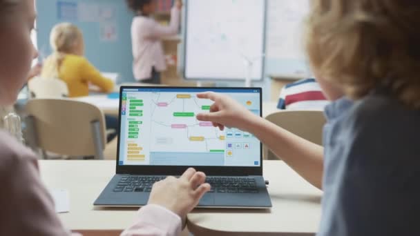 小学校理科の授業:肩の上に小さな男の子と女の子は、画面表示プログラミングソフトウェアとラップトップを使用します。物理学の先生は、スマートキッズがいっぱいの多様なクラスにレッスンを説明します — ストック動画