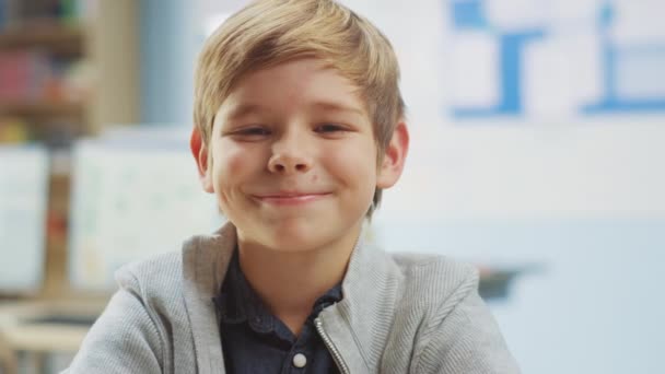 Retrato de um garotinho bonito sentado em sua mesa da escola, sorri feliz. Rapazinho esperto com sorriso encantador sentado na sala de aula. Close-up Camera Shot — Vídeo de Stock