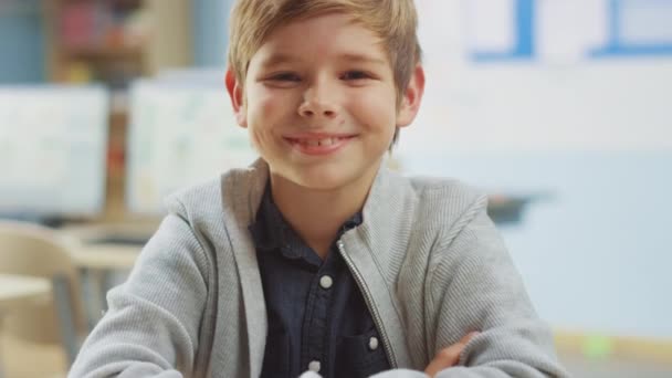Portret van een schattig jongetje dat bij zijn schoolbureau zit, lacht gelukkig. Slimme kleine jongen met een charmante glimlach zittend in de klaslokaal. Hefcamera neergeschoten. Langzame beweging — Stockvideo