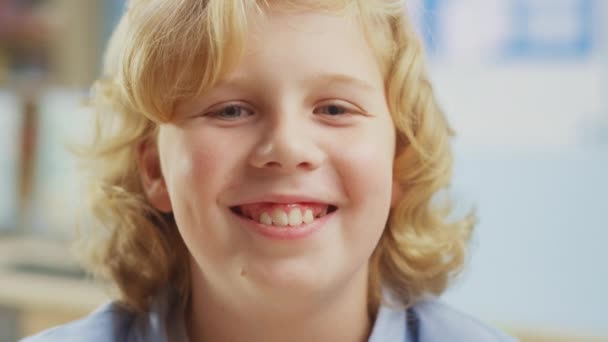 Портрет милого маленького мальчика с кудрявыми светлыми волосами, сидящего за школьным столом, улыбается счастливо. Умный мальчик с очаровательной улыбкой сидит в классе. Съемка крупным планом — стоковое видео