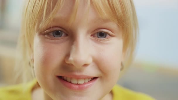 Retrato de una linda niña con el pelo rubio sentada en su escritorio de la escuela, sonríe felizmente. Niña inteligente con una sonrisa encantadora sentada en el aula. Primer plano de la cámara de tiro — Vídeos de Stock