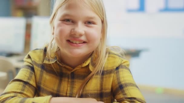 Portret van een schattig klein meisje met blond haar zittend aan haar schoolbureau, glimlacht gelukkig. Slimme kleine meid met een charmante glimlach zittend in de klaslokaal. Close-up camera neergeschoten — Stockvideo
